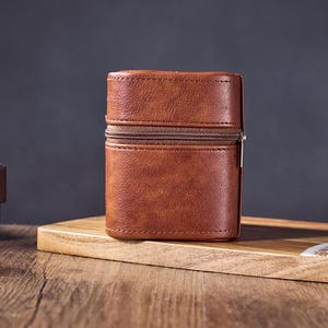 May include: A brown leather zippered case with a silver zipper pull. The case is sitting on a wooden surface.
