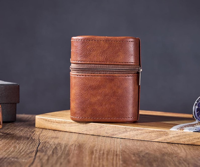 May include: A brown leather zippered case with a silver zipper pull. The case is sitting on a wooden surface.