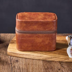 May include: A brown leather watch box with a zipper closure. The box is sitting on a wooden surface.