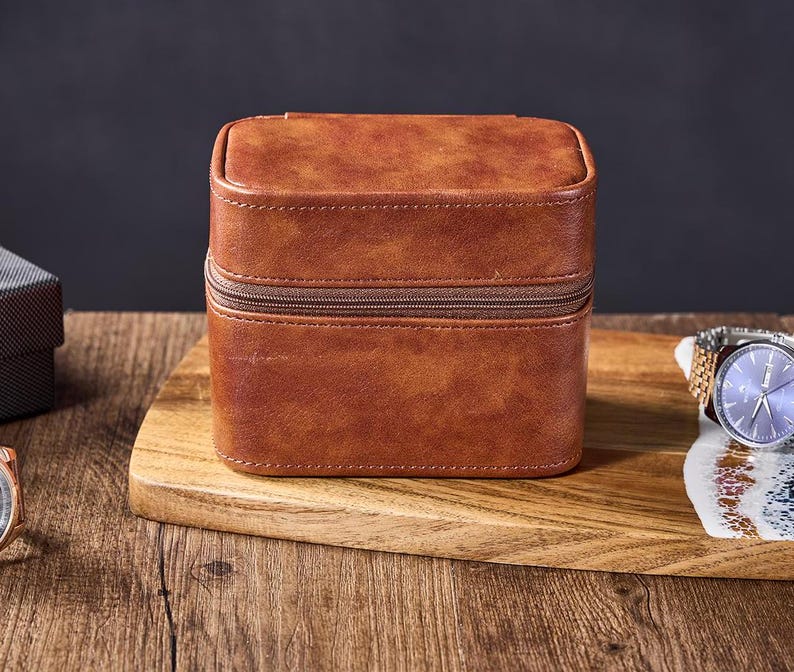 May include: A brown leather watch box with a zipper closure. The box is sitting on a wooden surface.