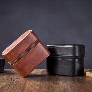 May include: Two leather watch boxes, one brown and one black, are sitting on a wooden surface. A watch is sitting on a wooden surface to the right of the boxes.