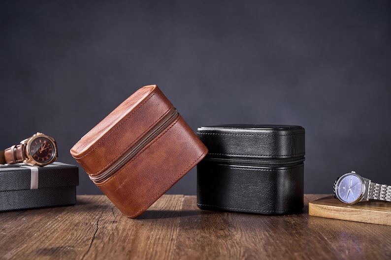 May include: Two leather watch boxes, one brown and one black, are sitting on a wooden surface. A watch is sitting on a wooden surface to the right of the boxes.
