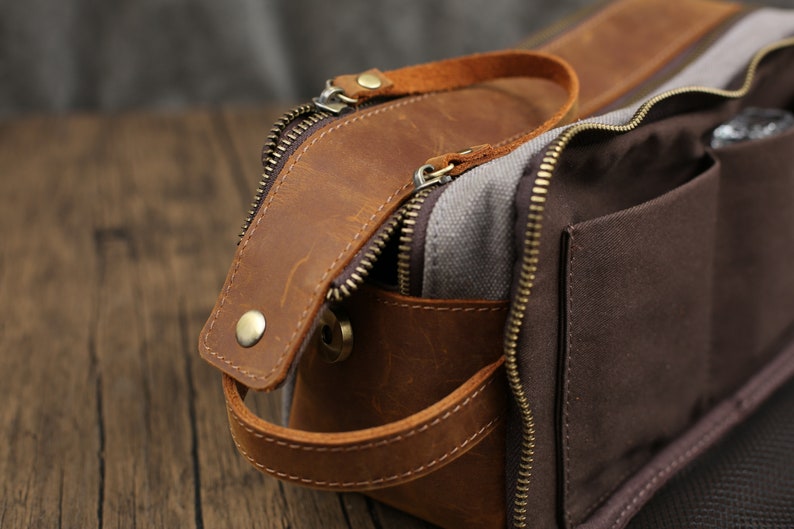 May include: A brown leather and canvas toiletry bag with a zipper closure and a handle. The bag has a brown leather exterior with a canvas interior and a brown zipper. The bag has a handle on the side.
