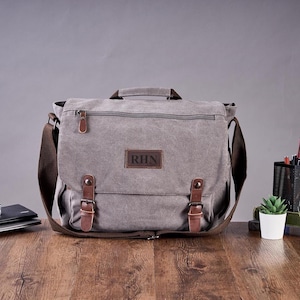 May include: A grey canvas messenger bag featuring a top handle, a brown leather patch with the letters RHN, and two brown leather straps with metal buckles. The bag includes a zippered front pocket and a wide brown adjustable shoulder strap.