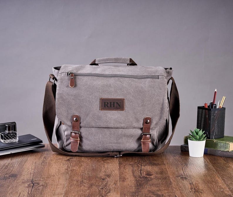 May include: A grey canvas messenger bag featuring a top handle, a brown leather patch with the letters RHN, and two brown leather straps with metal buckles. The bag includes a zippered front pocket and a wide brown adjustable shoulder strap.