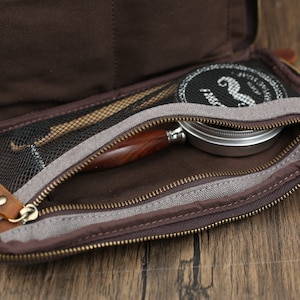 May include: Open brown leather tobacco pouch with a zippered compartment. Inside, a wooden-handled pipe, a tin, and tools are visible. The pouch has a mesh pocket and a brown interior. The zipper is gold-colored.
