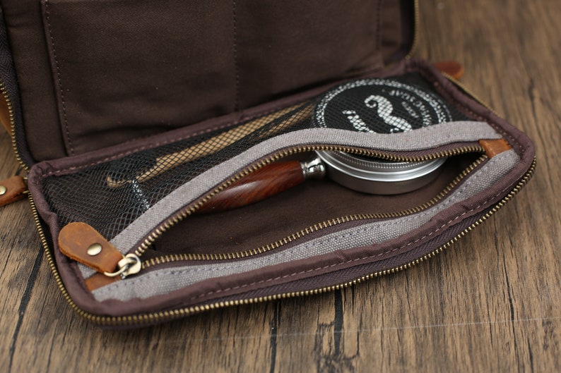 May include: Open brown leather tobacco pouch with a zippered compartment. Inside, a wooden-handled pipe, a tin, and tools are visible. The pouch has a mesh pocket and a brown interior. The zipper is gold-colored.