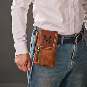 Puede incluir: Una funda de cinturón de cuero marrón con cierre de solapa y bolsillo con cremallera. La funda tiene un portalápices y presenta el nombre "MICHAEL" y los números romanos "IVI" en negro. La funda está sujeta a un cinturón negro que se lleva con vaqueros azules.