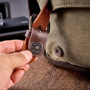 May include: An olive green canvas bag featuring a brown leather strap with a metal magnetic snap closure. The circular metal snap is engraved with text and secures the flap to the main body of the bag, which has a textured fabric surface.