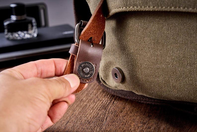 May include: An olive green canvas bag featuring a brown leather strap with a metal magnetic snap closure. The circular metal snap is engraved with text and secures the flap to the main body of the bag, which has a textured fabric surface.