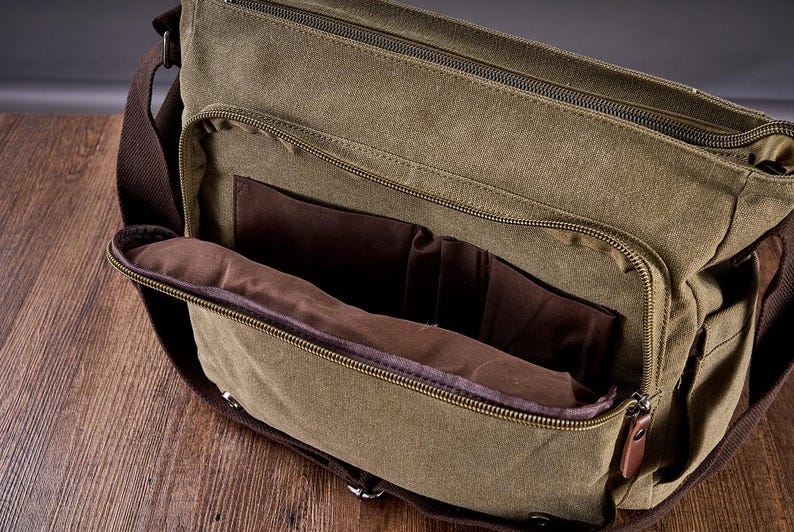 May include: An olive green canvas messenger bag featuring a large front zippered pocket that is partially open, revealing a dark brown fabric interior lining with internal organizational compartments and a brown adjustable shoulder strap.