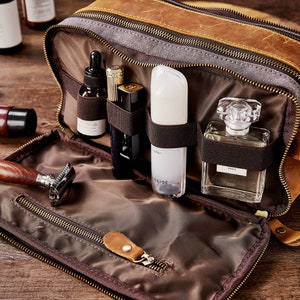 May include: A brown leather travel toiletry bag with a zippered opening. The bag is open and has several compartments for holding toiletries. The compartments are filled with various bottles and tubes of toiletries, including a razor, a bottle of perfume, and a bottle of lotion.