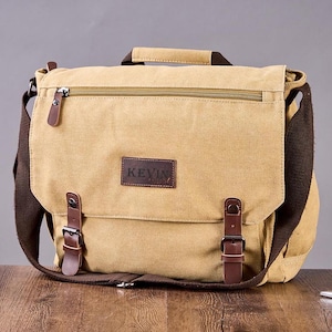 May include: A tan canvas messenger bag featuring a dark brown leather patch with the text KEVIN embossed on the front flap. The bag includes a top handle, a zippered pocket, and two brown leather straps with metal buckles for secure closure.
