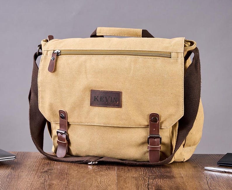May include: A tan canvas messenger bag featuring a dark brown leather patch with the text KEVIN embossed on the front flap. The bag includes a top handle, a zippered pocket, and two brown leather straps with metal buckles for secure closure.
