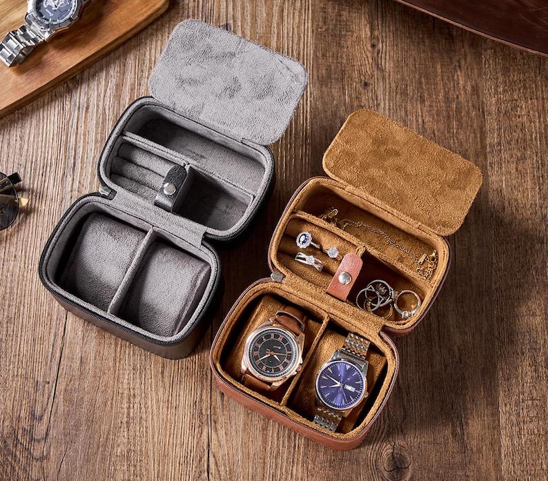 May include: Two leather watch and jewelry boxes. One box is brown with a gray interior and the other is black with a gray interior. Both boxes have compartments for watches and rings.