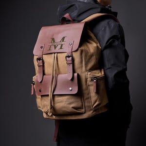May include: A brown canvas backpack with leather accents. The backpack has a large front pocket and a drawstring closure. The leather flap has the initial "M" and the name "MICHAEL" embossed in green.