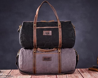 Può includere: Due borsoni in tela con dettagli in pelle. La borsa superiore è nera con una tracolla in pelle marrone e un logo a testa di cervo. La borsa inferiore è grigia con una tracolla in pelle marrone e le iniziali "RHW" su una toppa in pelle.