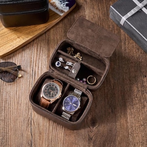 May include: A brown rectangular travel jewelry and watch case with a hinged lid. The interior features two padded watch cushions, a ring holder with three rings, a small compartment for jewelry, and a gold-tone snap closure tab.