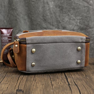 May include: A rectangular toiletry bag with a combination of gray canvas and brown leather. The bag features a top zipper, a leather handle, and brass-colored rivets on the bottom. The bag is designed for travel and personal care items.