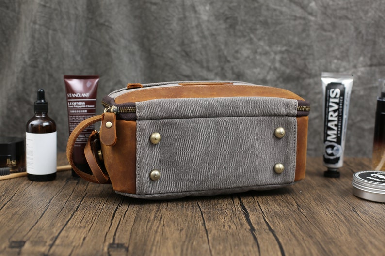 May include: A rectangular toiletry bag with a combination of gray canvas and brown leather. The bag features a top zipper, a leather handle, and brass-colored rivets on the bottom. The bag is designed for travel and personal care items.