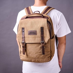 Può includere: Uno zaino in tela marrone con dettagli e fibbie in pelle. Lo zaino ha un ampio scomparto principale e una tasca anteriore più piccola. Lo zaino ha una toppa in pelle con il testo "RHK" sul davanti.