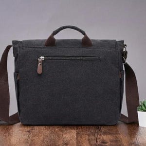 May include: A dark gray canvas messenger bag featuring a top handle, a brown adjustable shoulder strap, and a horizontal zippered pocket on the front with a brown leather-like pull tab. The bag has side pockets and a flap closure design.