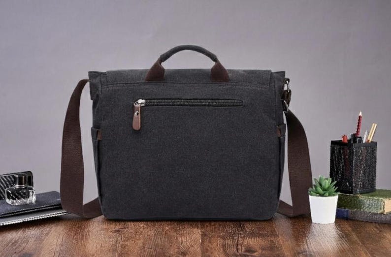 May include: A dark gray canvas messenger bag featuring a top handle, a brown adjustable shoulder strap, and a horizontal zippered pocket on the front with a brown leather-like pull tab. The bag has side pockets and a flap closure design.