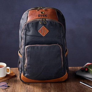 Puede incluir: Una mochila de lona negra con detalles de cuero marrón. La mochila tiene un compartimento principal grande, un bolsillo frontal más pequeño y dos bolsillos laterales. La mochila también tiene un parche de cuero con las iniciales "RHK" en la parte delantera.