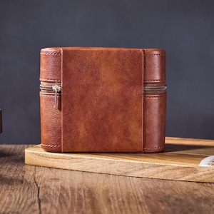 May include: A brown leather watch box with a zipper closure. The box is sitting on a wooden surface.
