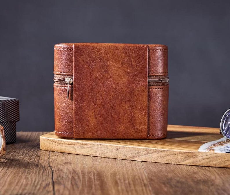 May include: A brown leather watch box with a zipper closure. The box is sitting on a wooden surface.