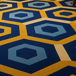 May include: A geometric patterned rug featuring a repeating hexagon design in navy blue, light blue, and gold. The rug is placed on a wooden floor, with a small black table and blue chair visible in the background.