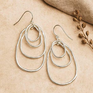 May include: A pair of silver-toned drop earrings featuring three interconnected, irregular oval-shaped wire loops of varying sizes. Each earring is suspended from a curved hook wire, showcasing a polished metallic finish and a layered design.