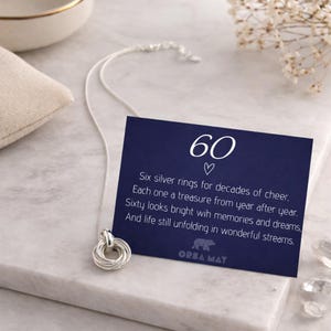 May include: A silver necklace featuring six intertwined rings, presented on a marble surface. A navy blue card with the number "60" and a poem about the rings is next to the necklace. The card also has the text "ORBA MAT".