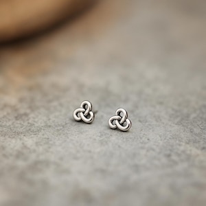 May include: Two small, silver-colored stud earrings. Each earring features a three-looped knot design. The earrings are displayed on a gray surface, with a blurred background.