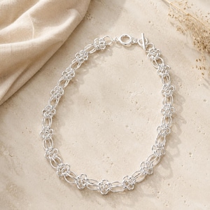 Può includere: Una collana a catena color argento caratterizzata da un motivo ripetitivo di maglie circolari interconnesse e motivi metallici a forma di fiore. La collana è fissata con una chiusura a T composta da un anello circolare e una barretta.
