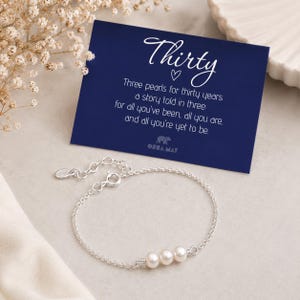 Op de afbeelding: Een zilveren kettingarmband met drie kleine witte parels, vergezeld van een donkerblauwe rechthoekige kaart met witte tekst: Thirty, Three pearls for thirty years, a story told in three: for all you've been, all you are, and all you're yet to be.