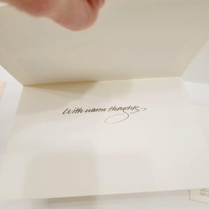 Op de afbeelding: Een open wenskaart met een cr&egrave;mekleurig interieur. De woorden "With warm thoughts" zijn geschreven in elegant zwart schrift. De kaart is opengeklapt en onthult de boodschap.