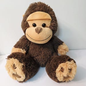 May include: A plush monkey toy with brown fur and tan accents on the face, hands, and feet. The monkey has a smiling expression with black button eyes and a tan nose. The toy is designed to sit upright.