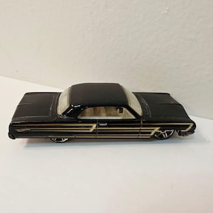 Può includere: Un'auto giocattolo in metallo pressofuso nera con dettagli a strisce dorate sui lati, interni bianchi e ruote color argento. Il veicolo presenta una carrozzeria dal profilo basso con un logo Hot Wheels visibile sul pannello laterale posteriore.