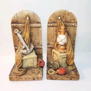 May include: Pair of nautical-themed bookends. Each bookend features a wooden door design with a weathered appearance. One has an anchor and fishing net, the other a lantern. Additional details include a life preserver and rope.