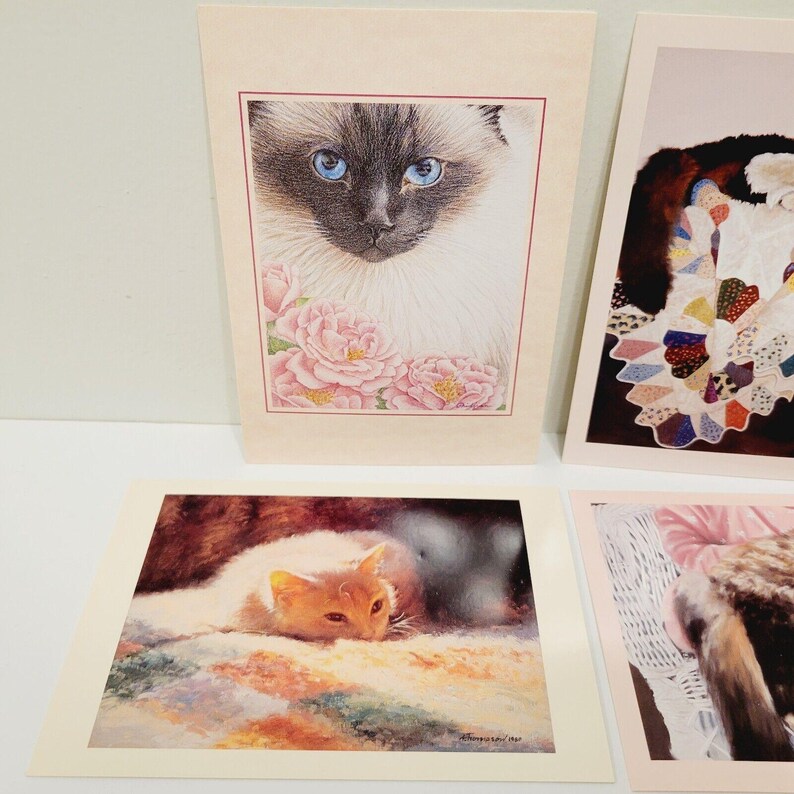 Op de afbeelding: Een verzameling kunstprints met katten als thema. E&eacute;n print toont een Siamees met blauwe ogen en roze rozen. Een andere toont een slapende oranje kat. De prints hebben een cr&egrave;mekleurige rand en worden op een wit oppervlak weergegeven.