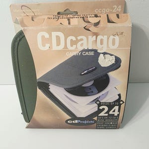 May include: An olive green CD cargo carry case in its original cardboard packaging. The box features the text CDcargo, cdProjects, and states it holds up to 24 CDs or 12 CDs with booklets. The packaging shows a gray zippered case.