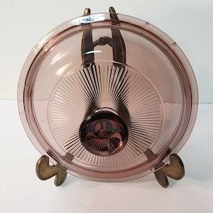 May include: A vintage, round, light purple glass lid with a dark purple knob. The lid has a sunburst design in the center and is displayed on a brass-colored metal stand. The lid is likely for a casserole dish.