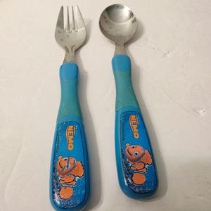 Może przedstawiać: Zestaw widelca i łyżki ze stali nierdzewnej z niebieskimi plastikowymi uchwytami. Każdy uchwyt zawiera logo Finding Nemo oraz grafikę pomarańczowej rybki Nemo. Sztućce mają metaliczne wykończenie oraz niebieskie elementy chwytne.