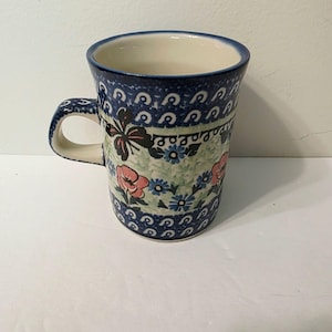 Może przedstawiać: Ceramiczny kubek z kremowym wnętrzem i ciemnoniebieskim brzegiem. Zewnętrzna strona ozdobiona jest powtarzającym się wzorem ciemnoniebieskich zawijasów u góry i u dołu oraz środkowym pasem z różowymi kwiatami, małymi niebieskimi kwiatkami i motylami.