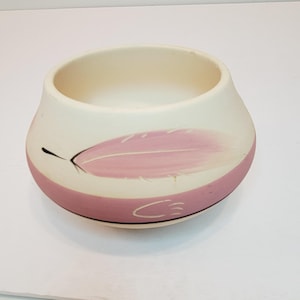 Puede incluir: Un pequeño cuenco de cerámica blanquecina con un diseño de pluma rosa. El cuenco tiene una banda rosa alrededor de la base y una línea negra en la parte inferior. La pluma está pintada en un color rosa suave.