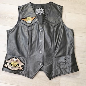 Może przedstawiać: Czarna skórzana kamizelka z dekoltem w serek i zapięciem na guziki. Posiada dwie naszywki: jedną z napisem "Ladies of Harley" i drugą z napisem "Harley Owners Group". Kamizelka ma metkę "First Genuine Leather".