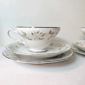 May include: White teacup and saucer set with a floral design and silver trim. The teacup has a curved handle and sits atop a matching saucer. The set includes a small plate, all with a delicate floral pattern.