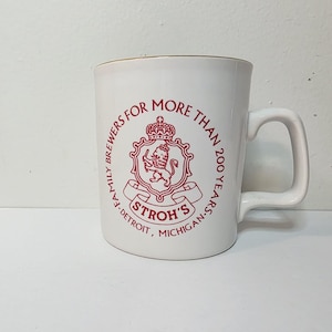 May include: White ceramic mug with a red Stroh's logo and text. The logo features a lion inside a crest with a crown. Text reads "Family Brewers for More Than 200 Years - Detroit, Michigan."