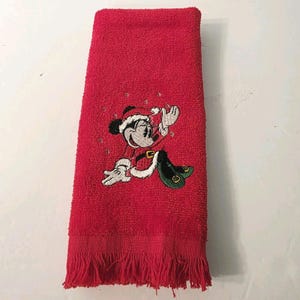 Puede incluir: Toalla de mano roja con una imagen bordada de Minnie Mouse con un traje de Pap&aacute; Noel. La toalla tiene un borde con flecos y est&aacute; hecha de un material suave y absorbente. El dise&ntilde;o de Minnie Mouse es en negro, blanco, rojo y verde.